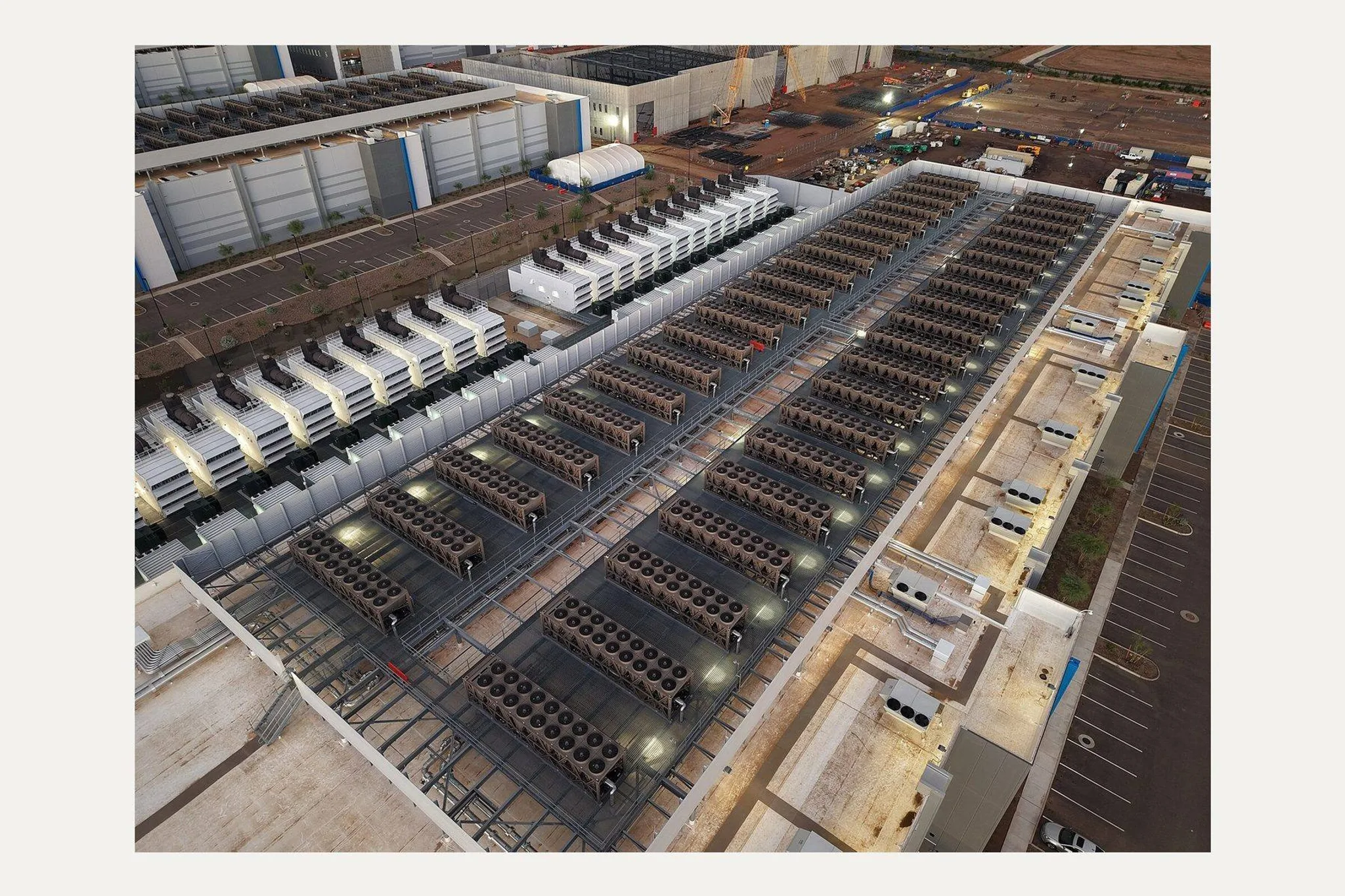 Aerial view at dusk of a data-centre roof lined with rows of fan-topped cooling units, with banks of backup generators alongside and construction cranes beyond.