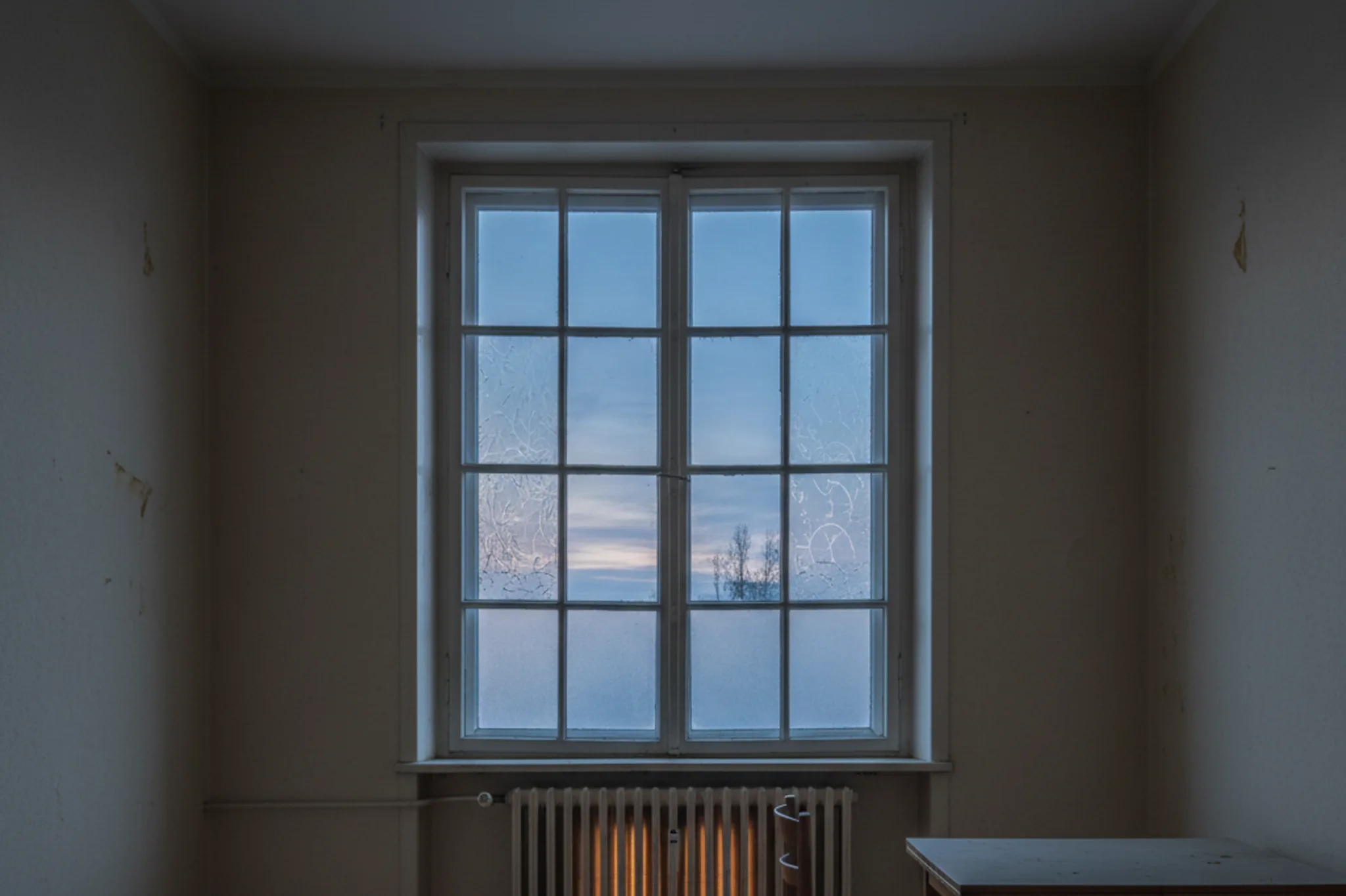 An empty European flat in winter dusk, a single warm radiator under a window, no one in the room